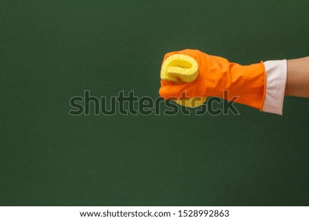 Woman's hand in orange protective glove with yellow sponge in a green background. Washing and cleaning concept.