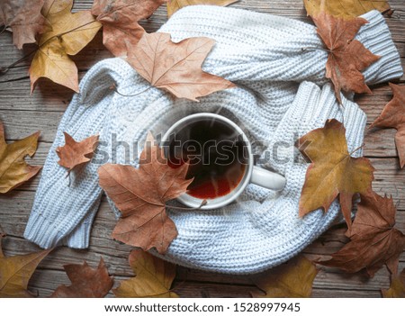 Cup of coffee wrapped in sweater on rustic wooden table with dry autumn leaves