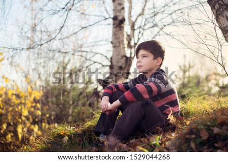 The boy sits on the grass under the trees and looks into the distance.