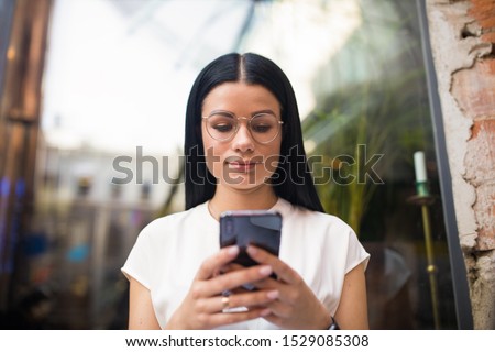 Stylish hipster girl in glasses online payment via mobile phone while sitting against coffee shop window. Female in spectacles fashion blogger reading article on cellphone 