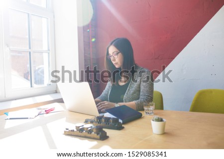 Female professional magazine editor typing text on web site via pc laptop computer while sitting in coworking space. Hipster girl in stylish glasses smart freelance worker using notebook for chat 