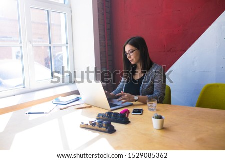 Female in casual wear and glasses experienced business plan writer browsing wifi via pc la[top computer while sitting in coworking office near window with copy space for advertising content 