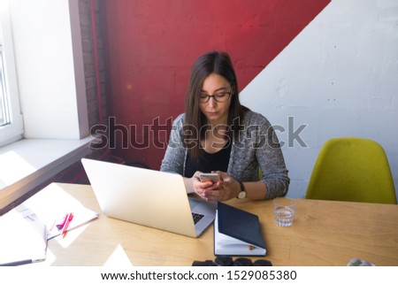 Business woman online payment via cell telephone while sitting at desktop with laptop computer during work day in office. Female freelancer received notifications on smartphone during work on netbook 