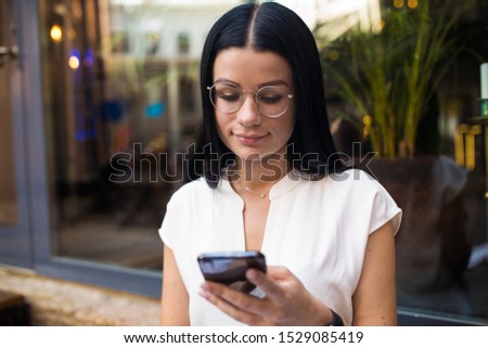 Charming dark hair woman in trendy glasses online shopping store via mobile phone during recreation time. Hipster girl in glasses using apps on cellphone 