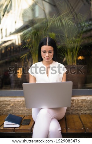 Business woman checking e-mail on laptop computer while sitting outdoors coffee shop during work break. Female in glasses successful freelance real estate analyst reading news via notebook gadget 