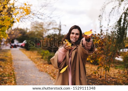 Cheerful young girl with long brown hair wearing autumn beige coat, walking at the park. Autumn walk. Woman portrait. Girl is playing and toss leaf and smiling. Woman enjoying fall nature. 