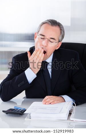 Mature Businessman Yawning While Calculating At Desk