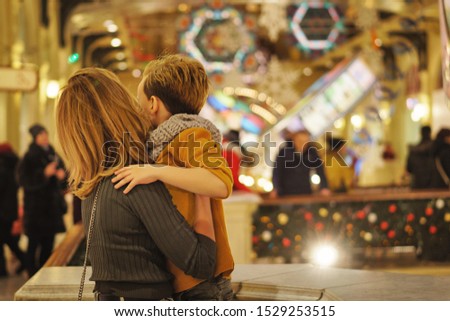 Dressed in stylish modern casual wear, mother and son in shopping mall decorated for Christmas. Garlands and christmas lights on background in defocus. Back view. Lifestyle concept. Copy space