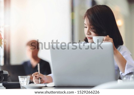Young businesswoman holding cup of coffee and writing with pen while working with laptop in modern office.