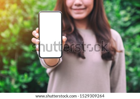 Mockup image of a woman holding and showing black mobile phone with blank screen with green nature background