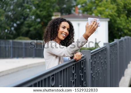 Cute young woman taking a selfie on a bridge with wrought iron railing posing for the camera on her smartphone