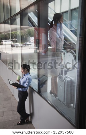 Young businesswoman inside the building talking on the phone, another women outside of the building looking at her notes