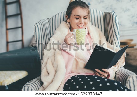 Cheerful attractive young woman sitting in chair and reading book. Also drink coffee or tea from cup and smile. Homie cozy look. Alone in room. Relaxed day Royalty-Free Stock Photo #1529378147