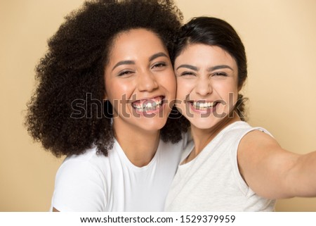 Excited multiethnic millennial girlfriends isolated on orange brown studio background make selfie on smartphone, happy overjoyed international young female friends take self-portrait picture together