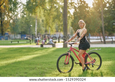 young woman in a short black summer dress rides a bicycle on a lawn with green grass in a city park