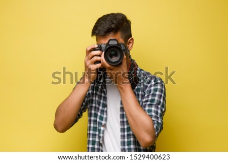 Handsome and confident indian man photographer with a large professional camera taking pictures photo shooting on the on the yellow background.
