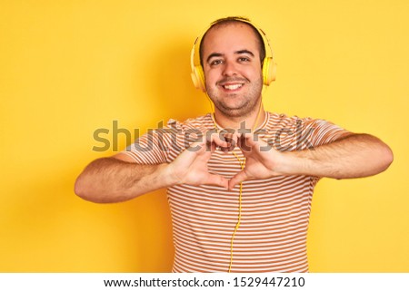 Young man listening to music using headphones standing over isolated yellow background smiling in love showing heart symbol and shape with hands. Romantic concept.