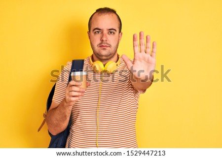 Student man wearing backpack headphones drinking coffee over isolated yellow background with open hand doing stop sign with serious and confident expression, defense gesture