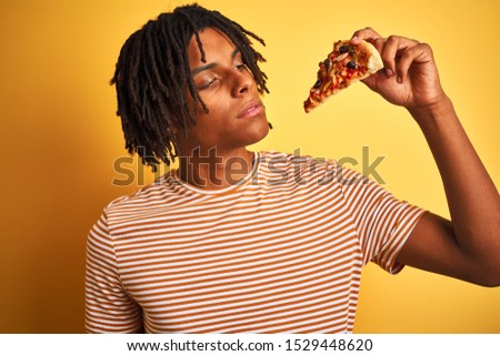 Afro american man with dreadlocks eating slice of pizza over isolated yellow background with a confident expression on smart face thinking serious