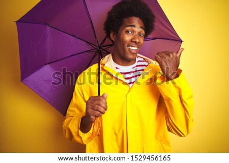 Afro american man wearing rain coat and umbrella standing over isolated yellow background pointing and showing with thumb up to the side with happy face smiling