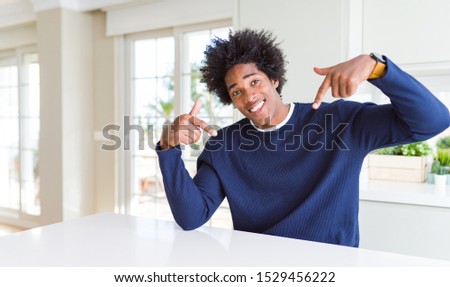 Young african american man wearing casual sweater sitting at home looking confident with smile on face, pointing oneself with fingers proud and happy.