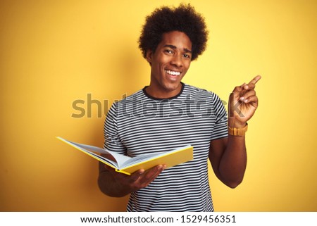 Afro american student man reading book standing over isolated yellow background very happy pointing with hand and finger to the side