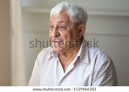 Elderly 70s man having hoary head positive mood toothy smile stands indoors looking out the window enjoy life and sunny day, concept of nursing home good professional care, healthy strong old age male