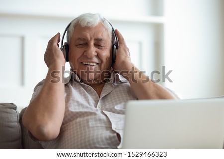 Senior man seated on sofa in living room hold laptop on lap wearing headphones listens favourite track having nostalgic mood enjoys songs of his youth, older generation using modern technology concept Royalty-Free Stock Photo #1529466323