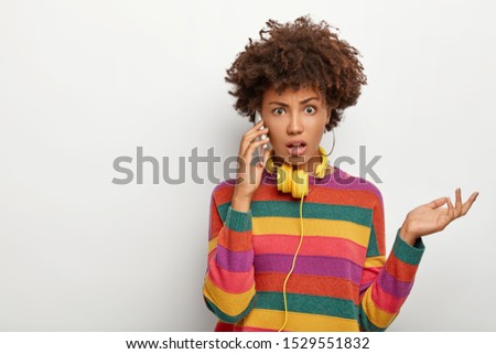 Photo of puzzled indignant African American woman raises palm, talks with friend via cellular, dressed in casual clothes, looks unhappy, poses against white background. Modern life and new technology