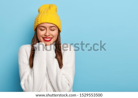 Happy Caucasian woman smiles pleasantly, has red painted lips, keeps hands under chin, wears cozy white winter sweater and yellow hat, keeps eyes closed, isolated over blue background, feels lucky
