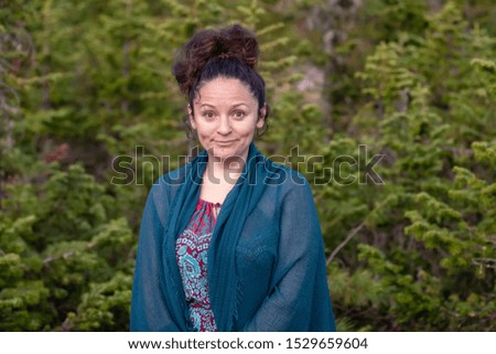 Portrait of a cute European girl with a surprised expression on her face with a blue scarf on her shoulders against the background of a coniferous blurred forest. Selective focus on the girl.