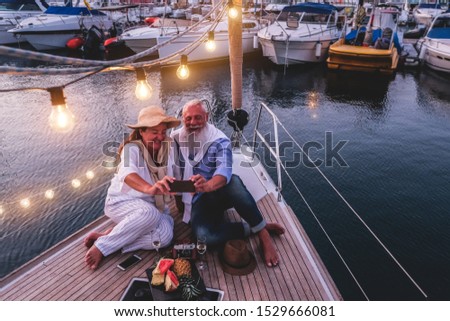 Happy senior couple taking selfie photo at night time on a sailboat, during anniversary vacation - Joyful elderly lifestyle, love, travel and holidays concept - Focus on hands phone