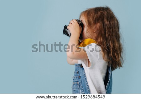 Toddler girl with curly hair making photo with retro camera isolated on blue wall half-face