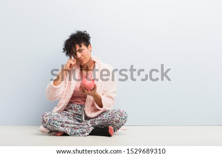 Young african american woman sitting with a piggy bank pointing his temple with finger, thinking, focused on a task.