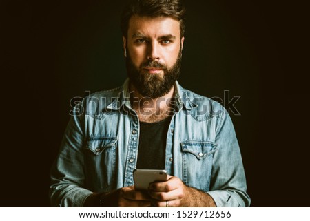 Portrait of young bearded man with smartphone in hand. Hipster guy in denim jacket using mobile phone for online communication in social media, networks. Male person texting messages, checking updates