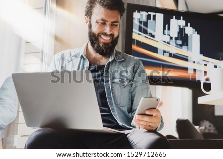 Male professional working on laptop and smartphone in office. Digital developer testing apps and synchronizing corporate programs. Manager taking selfie at workplace and posting images in social media