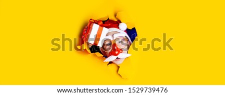 little cheerful Santa in glasses with a red nose and mustache gives a gift, getting out of the ragged yellow background