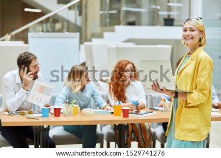 Positive delighted blonde woman, wears colourful outfit, keeps laptop computer in hands, turns head at camera with broad lovely smile, looks satisfied, expresses happiness on background of colleagues