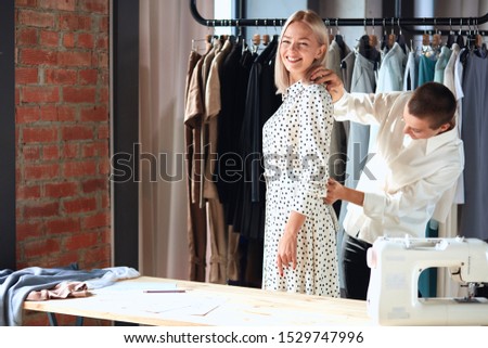 Modern and happy caucasian designer happily takes measurements of young female in room. Background clothes