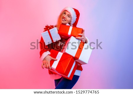 Happy excited young woman wearing Santa hat holding many boxes standing in background with red and blue neon light
