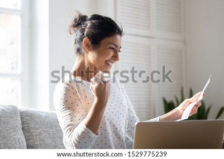 Happy Indian woman reading good news in letter, notification, holding paper, smiling girl rejoicing success, student excited by positive exam results, showing yes gesture, sitting on couch at home Royalty-Free Stock Photo #1529775239