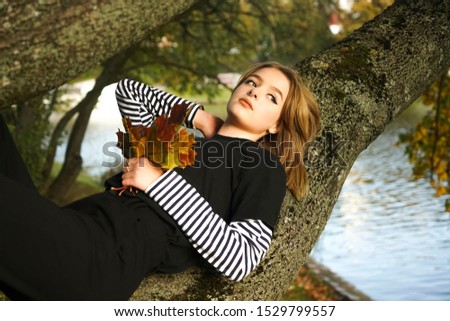 Beautiful young adult girl wear black dress and relax on tree trunk and relaxing deep into autumn fall forest. Cute blond woman enjoy nice day in autumn park. Empty copy space for inscription