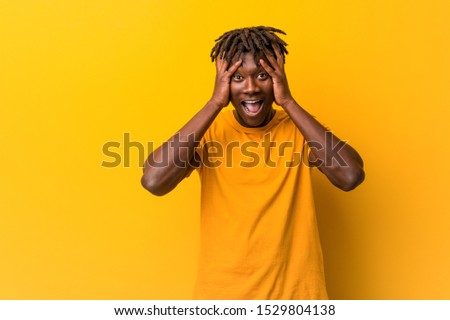 Young black man wearing rastas over yellow background laughs joyfully keeping hands on head. Happiness concept.