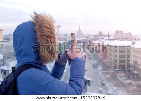 Unrecognizable woman tourist traveler in down jacket shoots video on smartphone on observation deck in city on roof of building, side view. Tourism, travel and journey concept. Moscow city in winter.