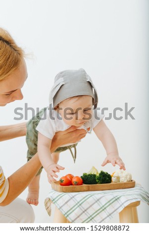 Active restless infant child with rabbit ears hat is reaching for vegetables while mother holds him in air.