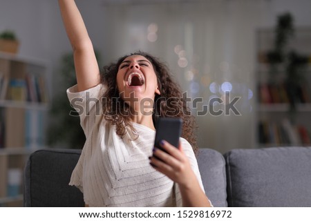 Excited woman celebrating success holding mobile phone sitting on a couch in the night at home Royalty-Free Stock Photo #1529816972