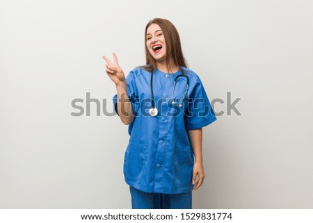 Young nurse woman against a white wall joyful and carefree showing a peace symbol with fingers.