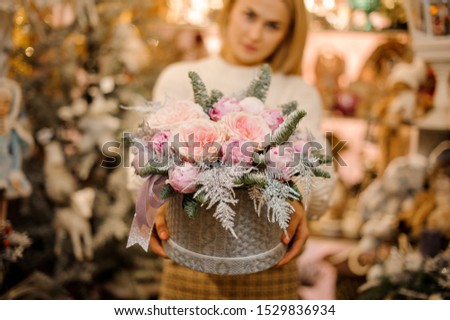 Woman holding a grey sweater pattern box with light pink peony roses decorated with fir-tree branches in the blurred background of flower shop