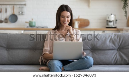 Smiling young woman using laptop, sitting on couch at home, beautiful girl shopping or chatting online in social network, having fun, watching movie, freelancer working on computer project Royalty-Free Stock Photo #1529846093