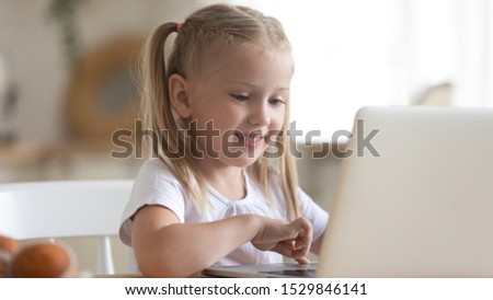 Smiling cute little girl using laptop close up, looking at screen, playing computer game, watching video or cartoons, sitting at table, having fun with gadget alone, electronic device addiction